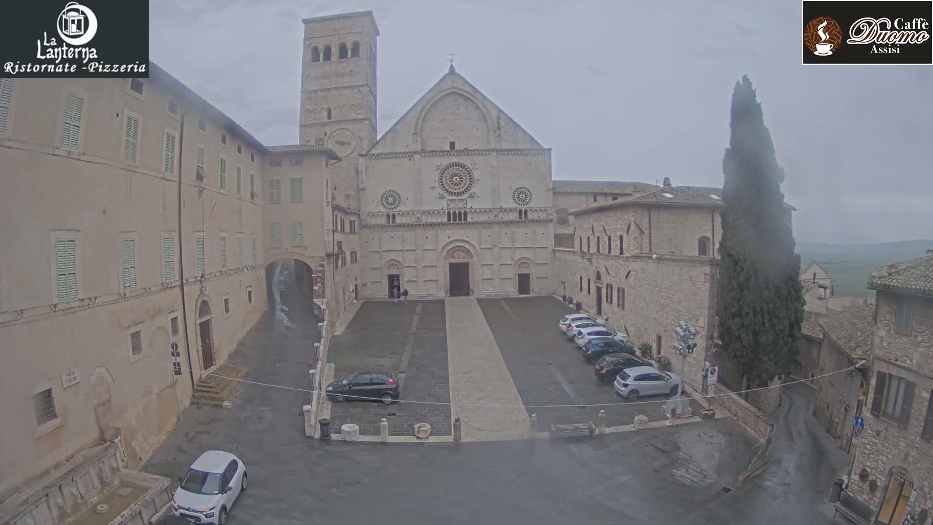 A large stone cathedral sits in a town square on an overcast day, with several cars parked nearby and light rain falling.