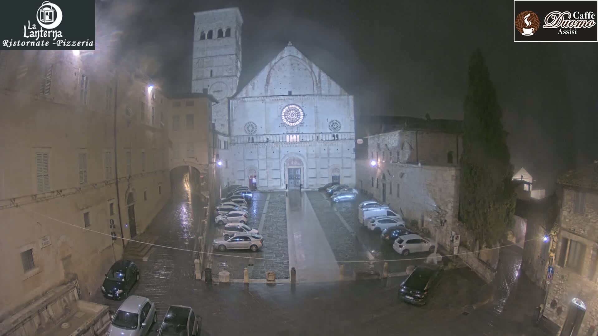 A large stone cathedral is surrounded by buildings and parked cars in a rain-soaked square at night.