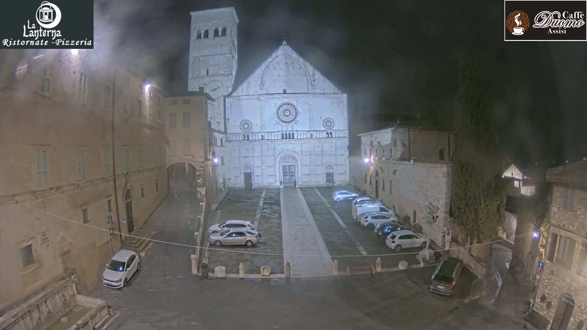 A large stone cathedral is situated in a town square at night, under a misty sky, with several cars parked in the square.