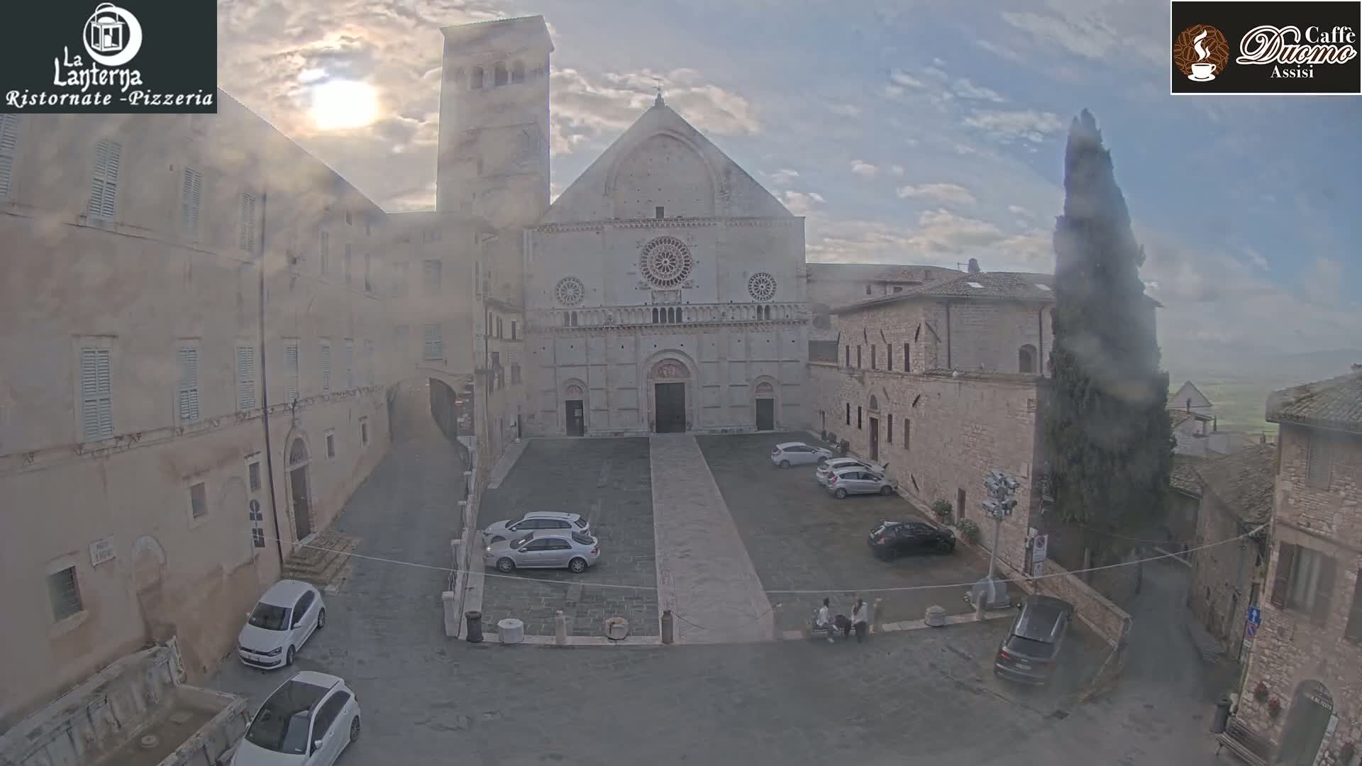 A hazy sun shines on a cathedral square in a historic town, where several cars are parked, and tall buildings surround a central plaza.
