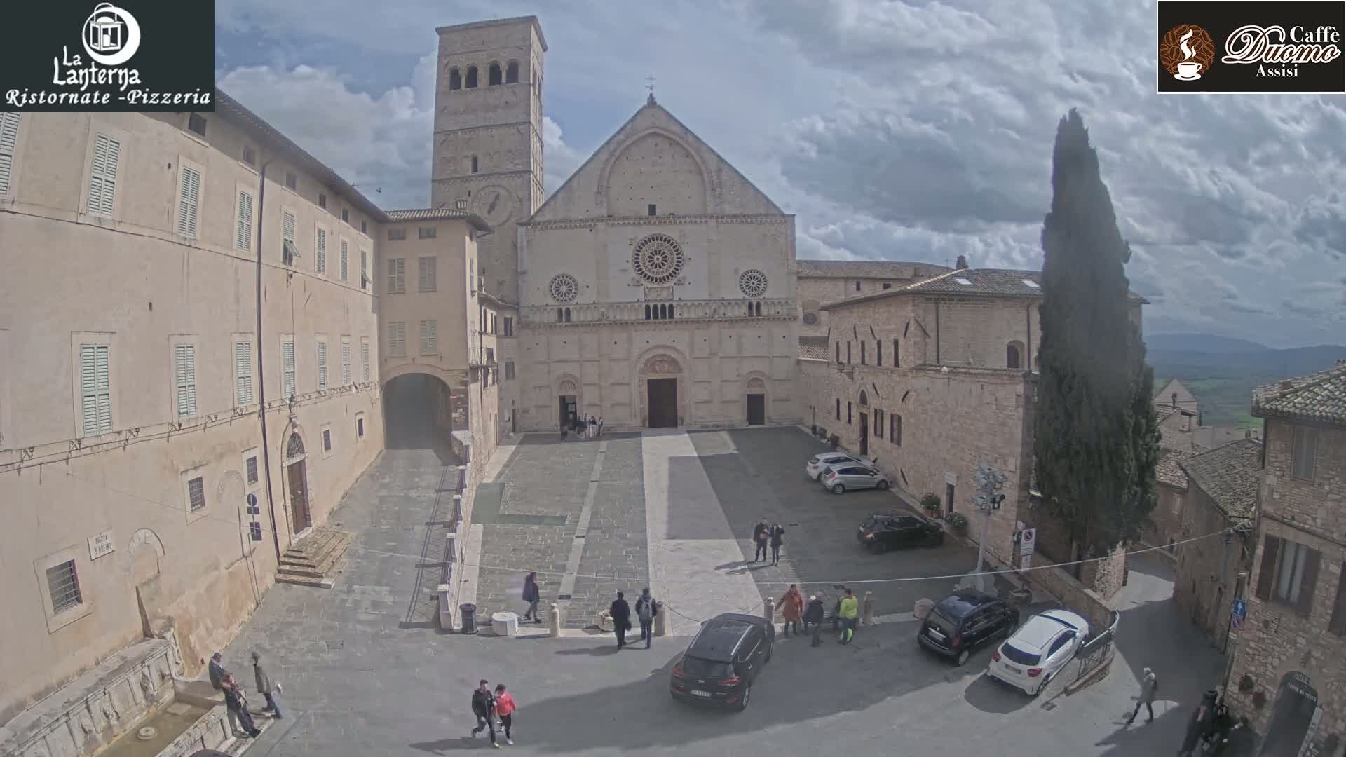 A stone plaza with a large cathedral in the background, surrounded by buildings under a partly cloudy sky.