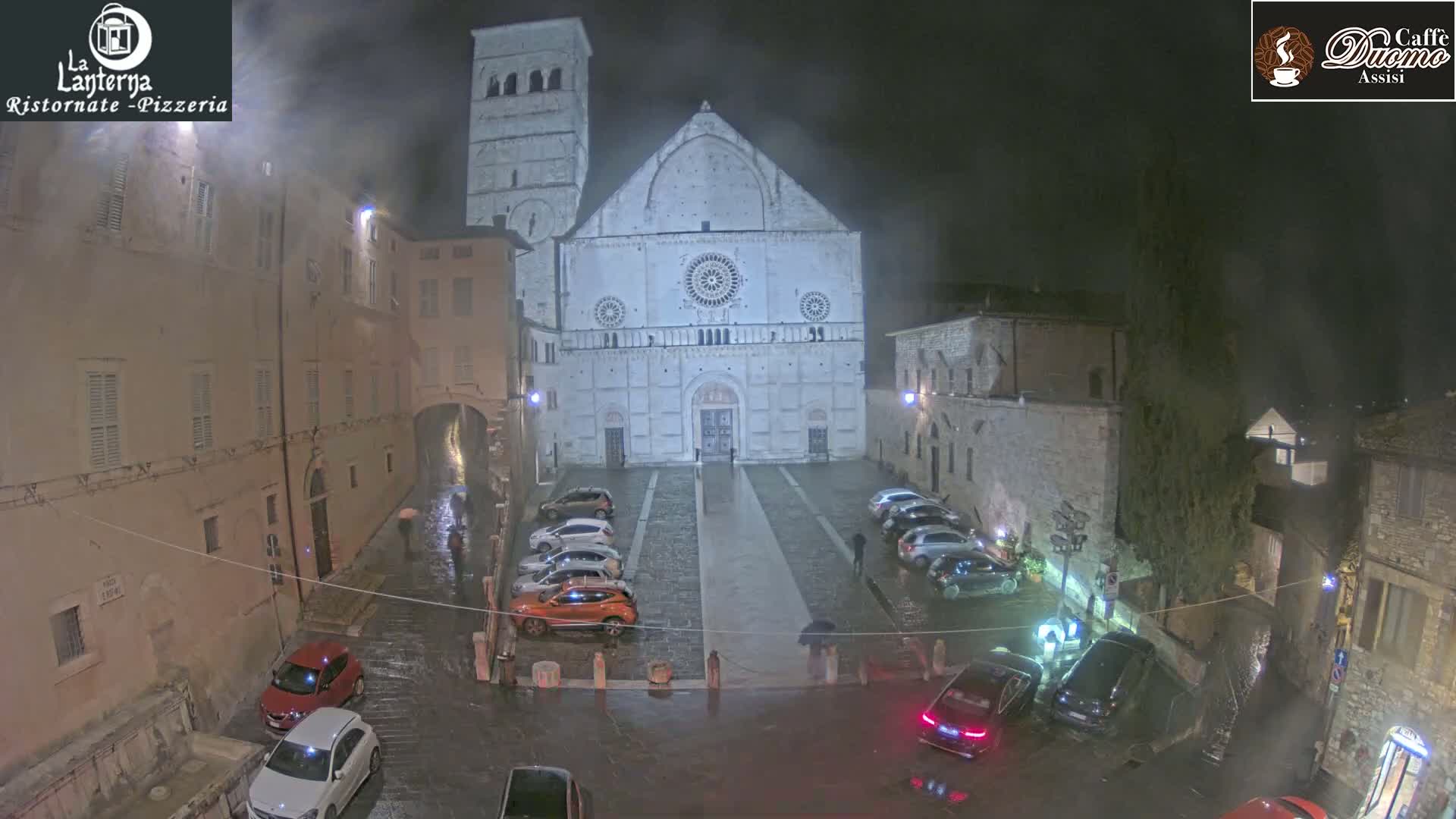 At night, under rainy conditions, a large stone cathedral is visible in a town square with cars parked around it, and buildings lining the square.