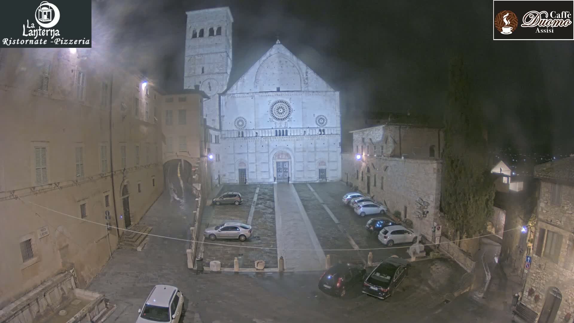 A nighttime view of a town square with cars parked in front of a large, light-colored cathedral under a hazy sky.