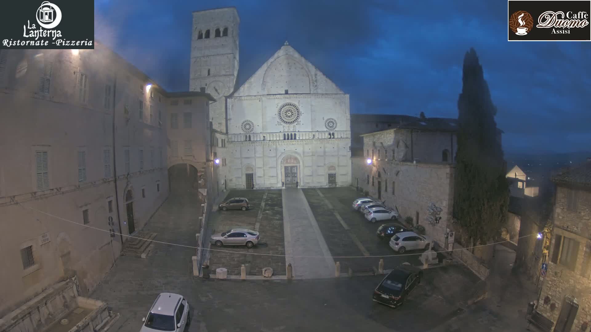 A large stone cathedral stands in a dimly lit, empty square at dusk, surrounded by older buildings, with several cars parked nearby.