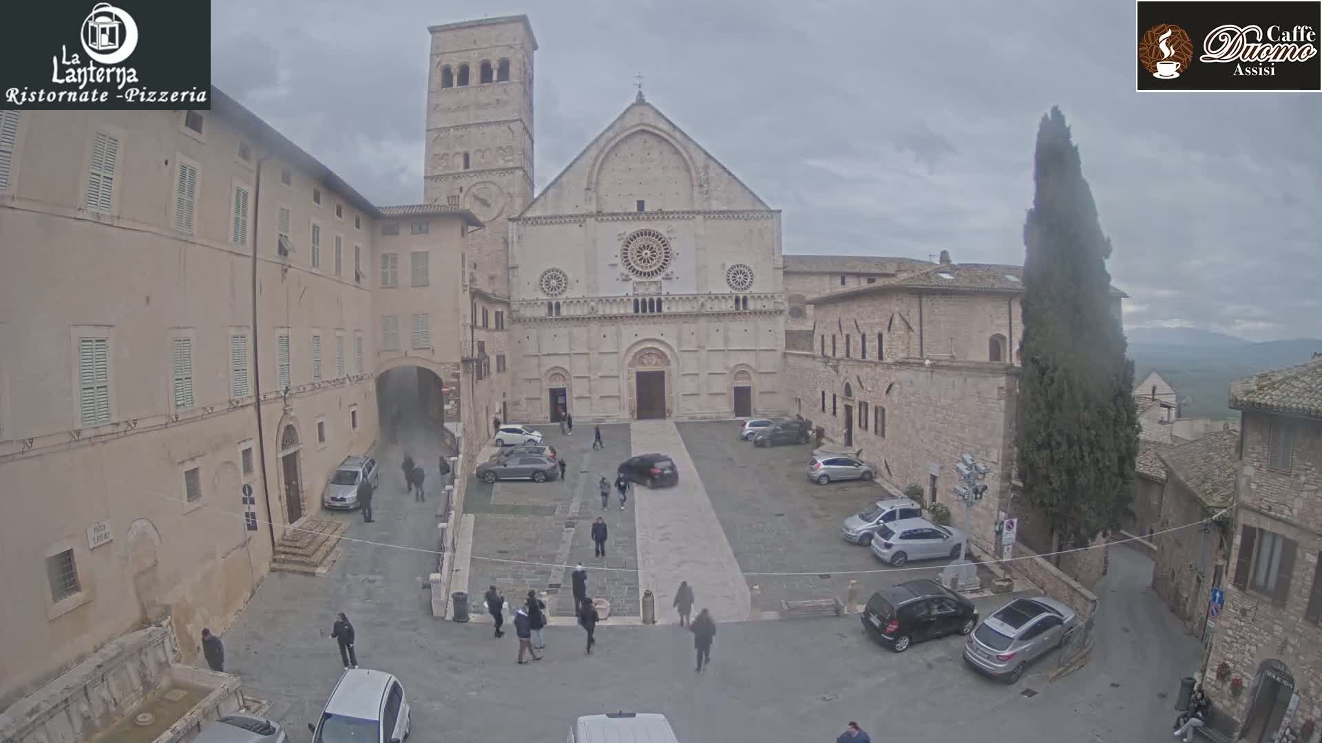 A stone cathedral and surrounding buildings are visible in a town square on an overcast day, with several cars parked and people walking around.