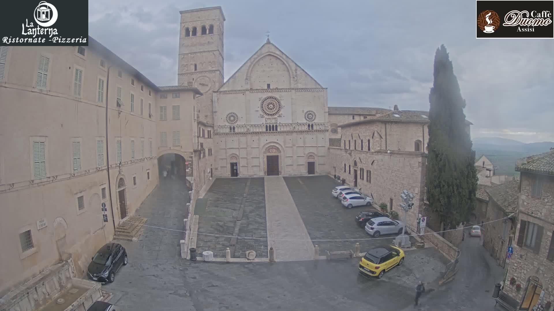 A large stone cathedral faces a plaza with several parked cars under an overcast sky.