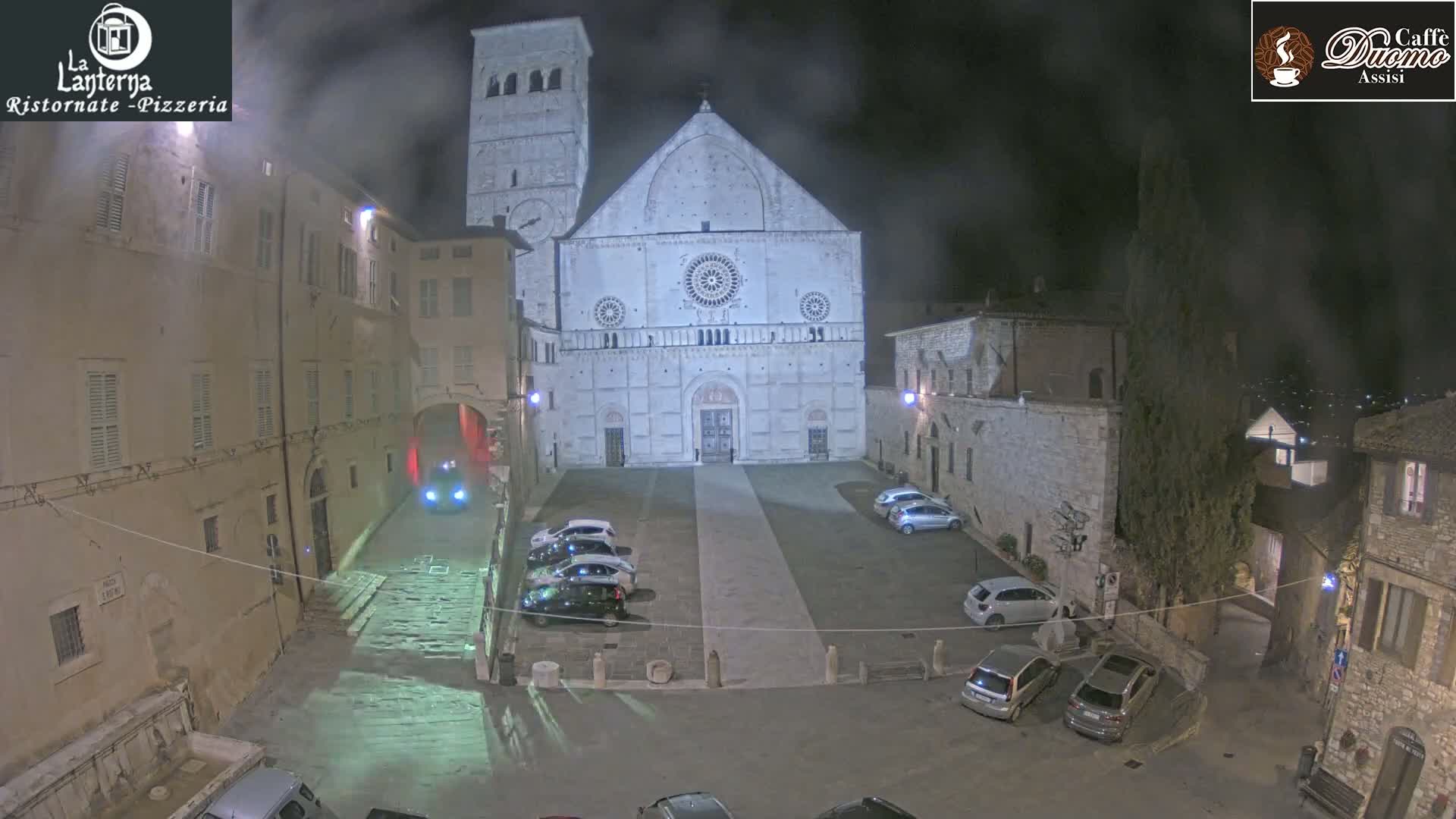 A nighttime view of a town square with a large cathedral and several parked cars under a hazy sky.