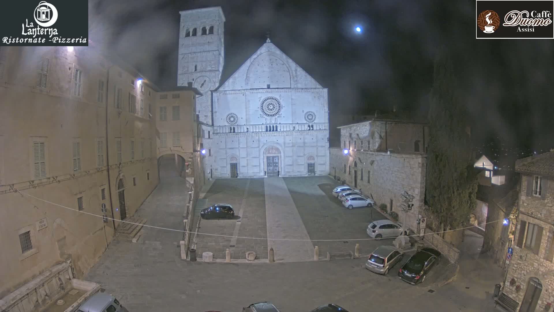 A nighttime view of a town square dominated by a large stone cathedral, under a hazy sky with a visible moon.