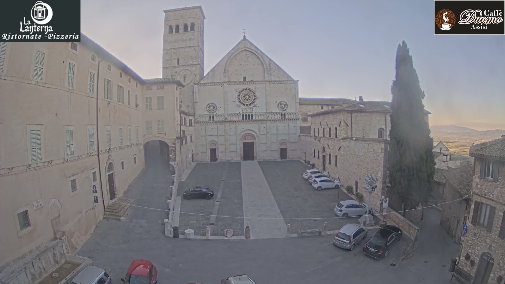 A large stone cathedral faces a plaza with several parked cars under a hazy, light sky.