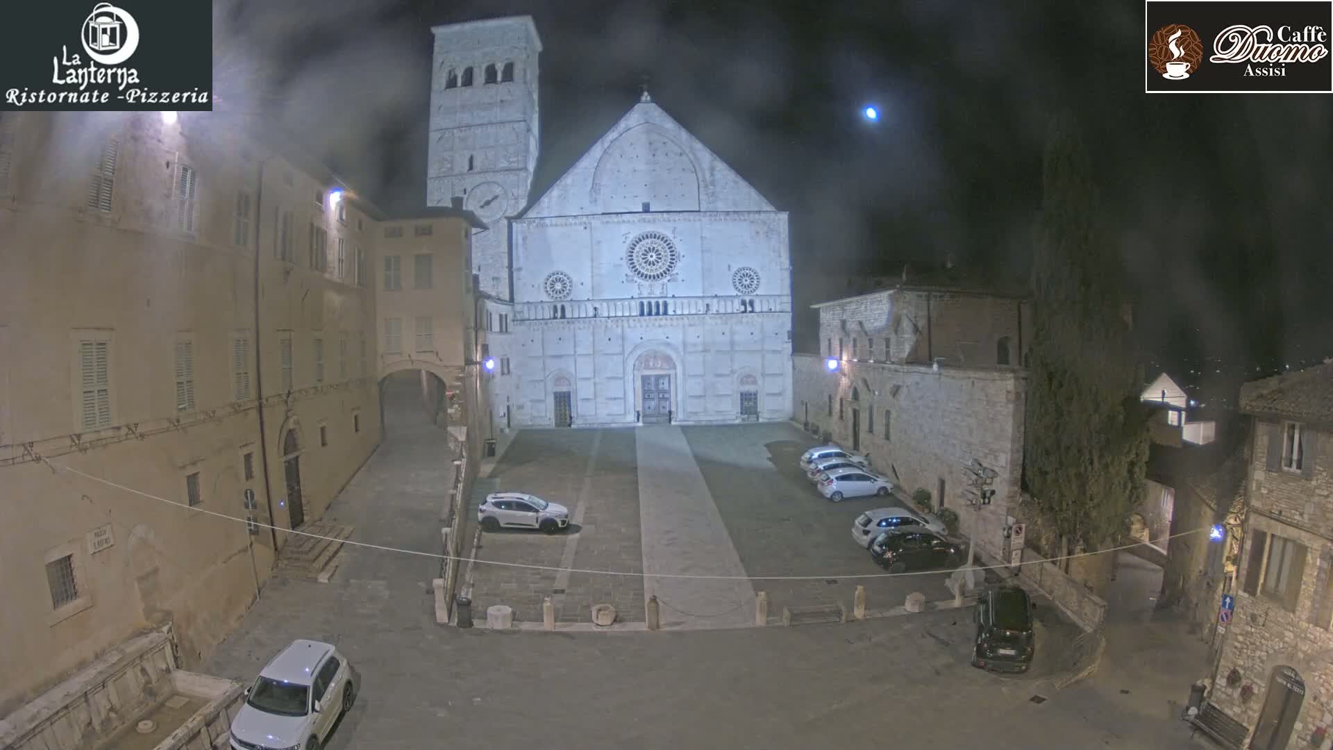 A large, light-colored cathedral stands in a dimly lit, stone plaza at night, surrounded by several parked cars and older buildings under hazy, cloudy skies.