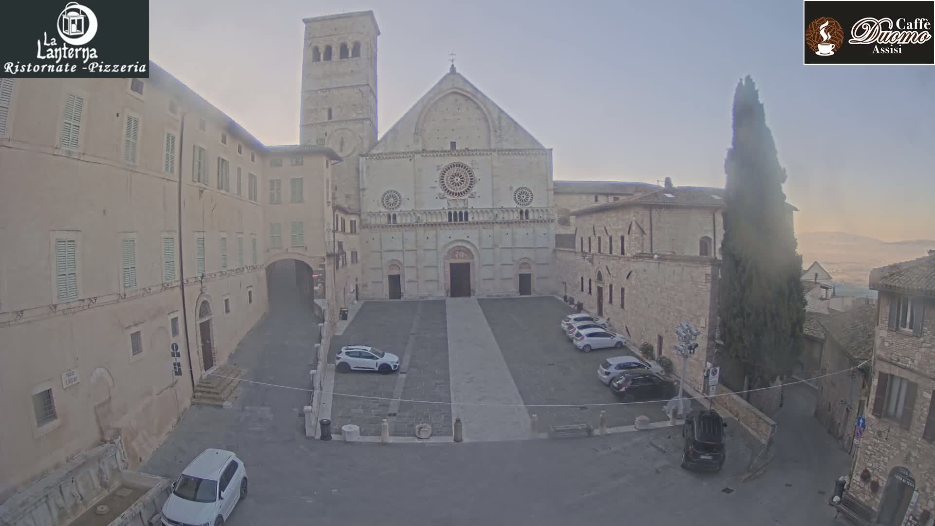 A large stone cathedral faces a town square with several parked cars, under a hazy, light sky.