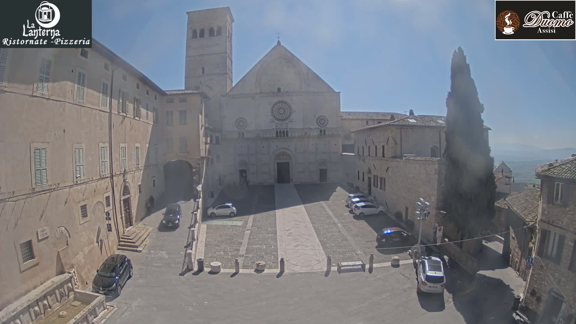 A large, light-colored stone church stands in a town square on a sunny day, surrounded by older stone buildings and several parked cars.