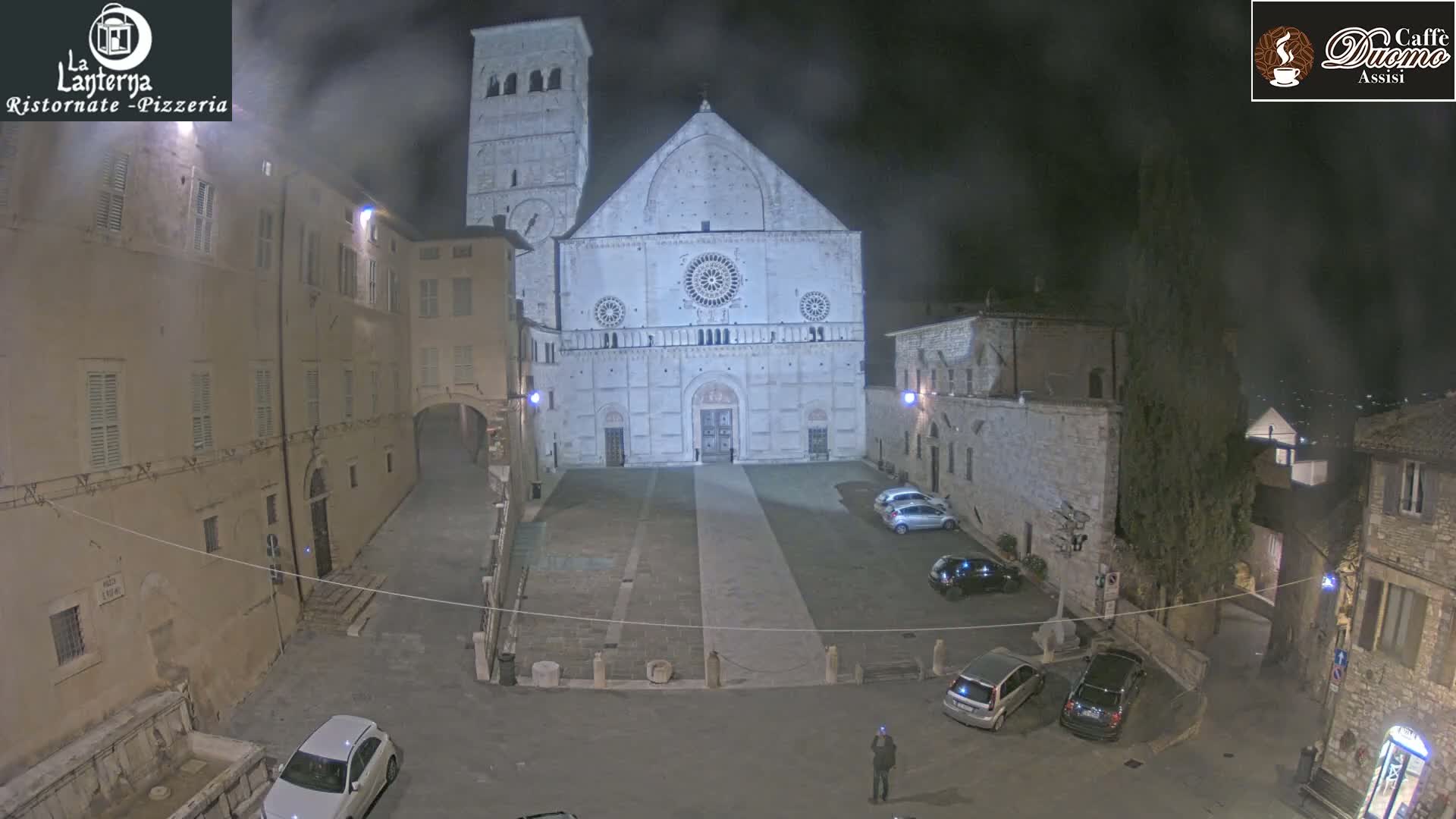 A nighttime view of a town square with a large stone cathedral at its center, under hazy or foggy conditions.