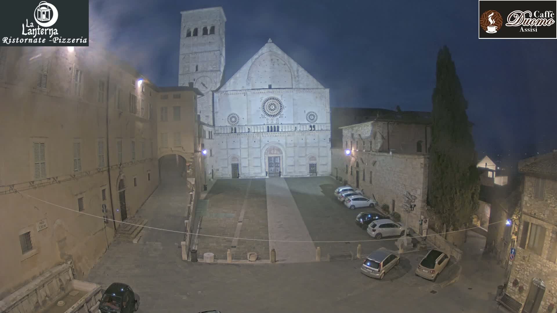 A nighttime view of a large cathedral in a town square, with several cars parked nearby and a slight haze in the air.