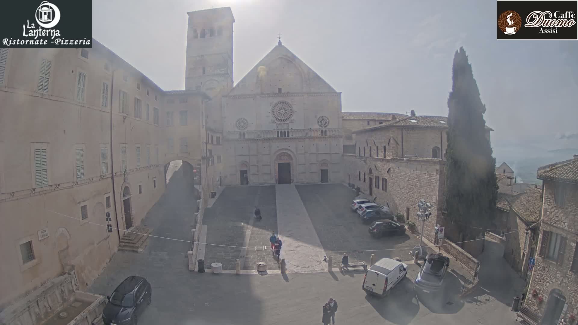 A hazy, sunny day reveals a large stone cathedral in a town square, surrounded by other stone buildings and a few parked cars.