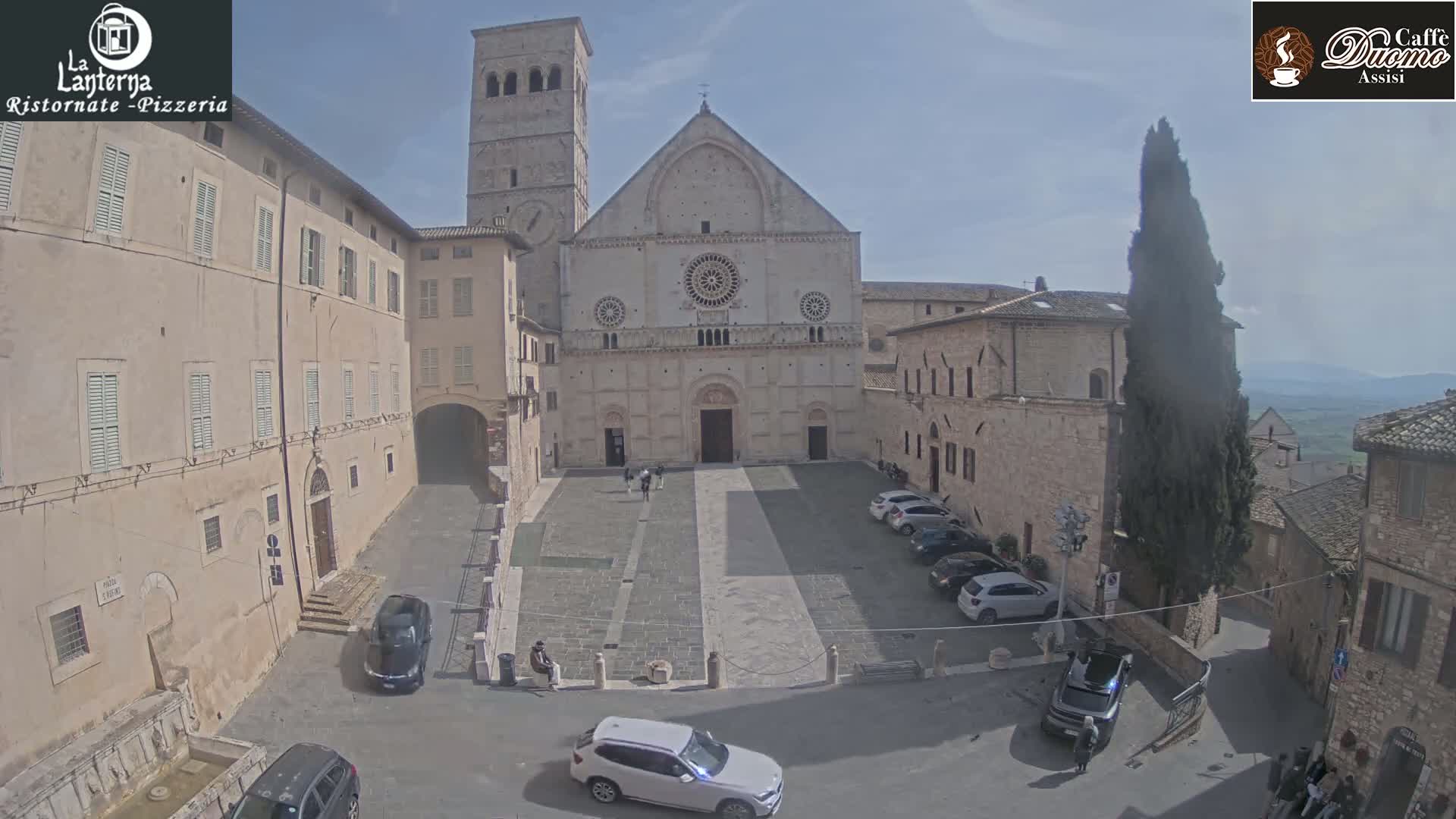 A large stone cathedral sits in a town square surrounded by older buildings, under a mostly sunny sky.
