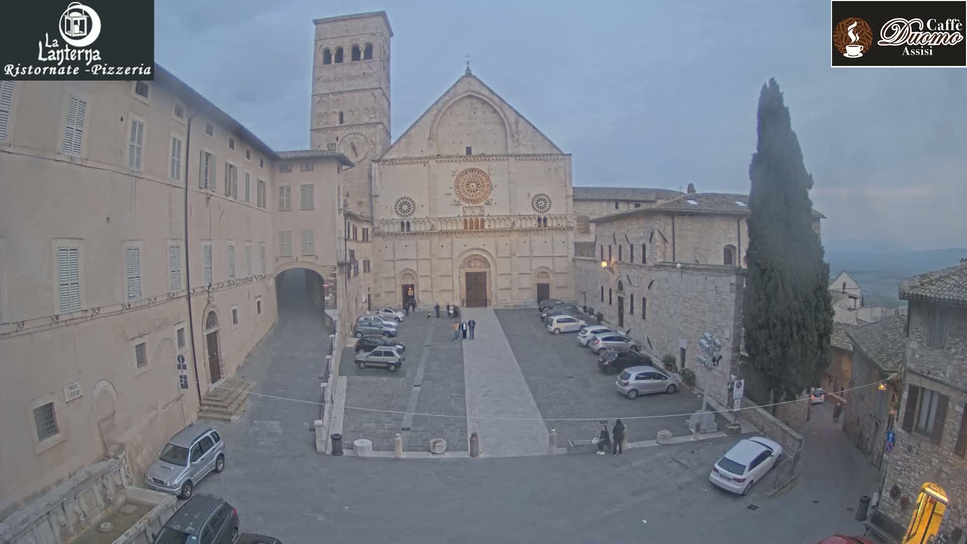 A large stone cathedral sits in a town square with several cars parked around it under an overcast sky.