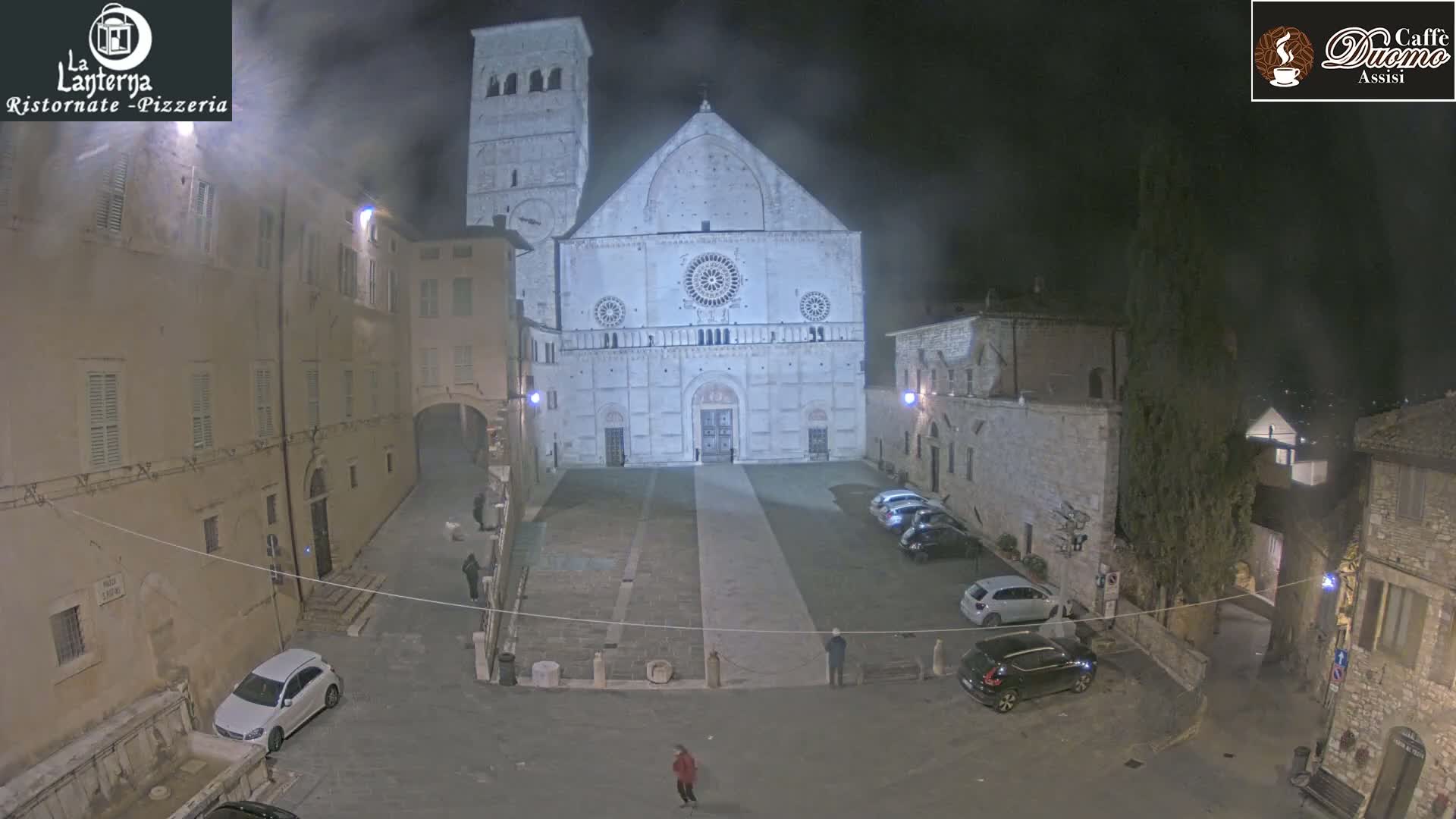 A nighttime view of a town square, featuring a large stone cathedral and several cars parked in the square under a hazy, dark sky.