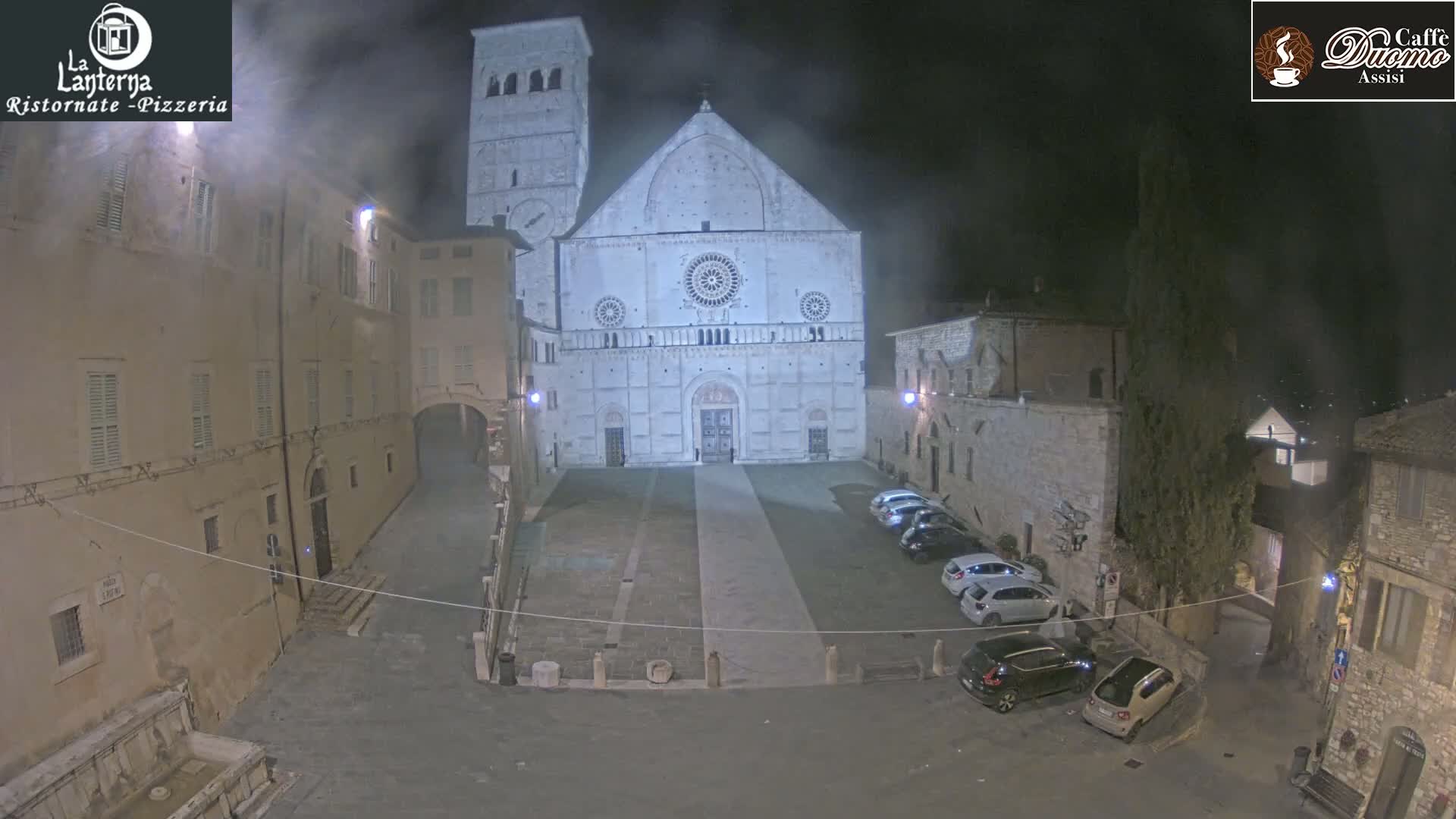 A nighttime view of a town square with a large cathedral, several parked cars, and buildings illuminated by artificial light in foggy conditions.