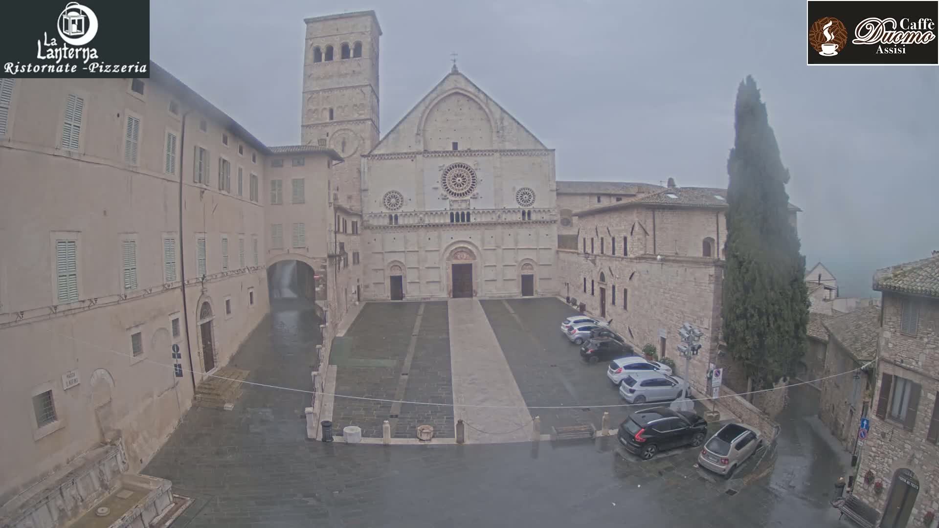 A large cathedral sits in a town square, surrounded by stone buildings in the rain.