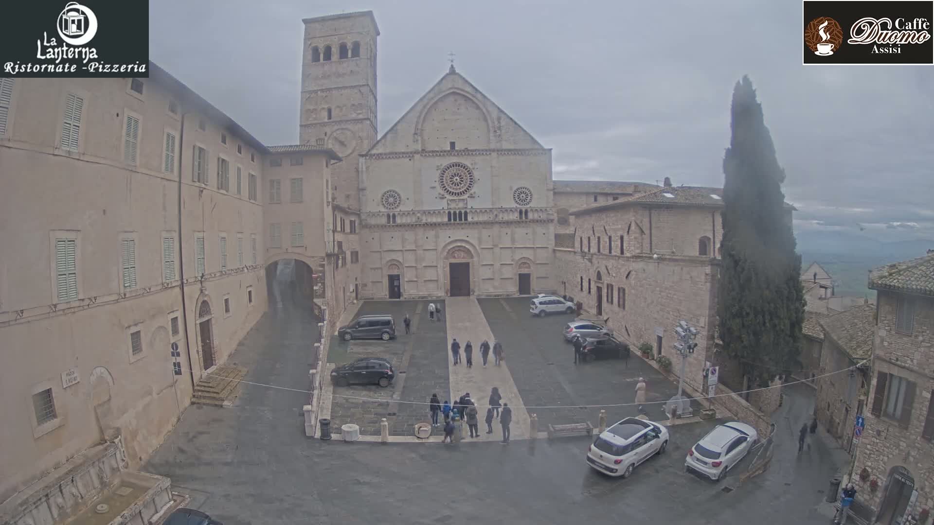 A large stone cathedral is situated in a town square, surrounded by buildings, with several cars parked and people walking around under an overcast sky.