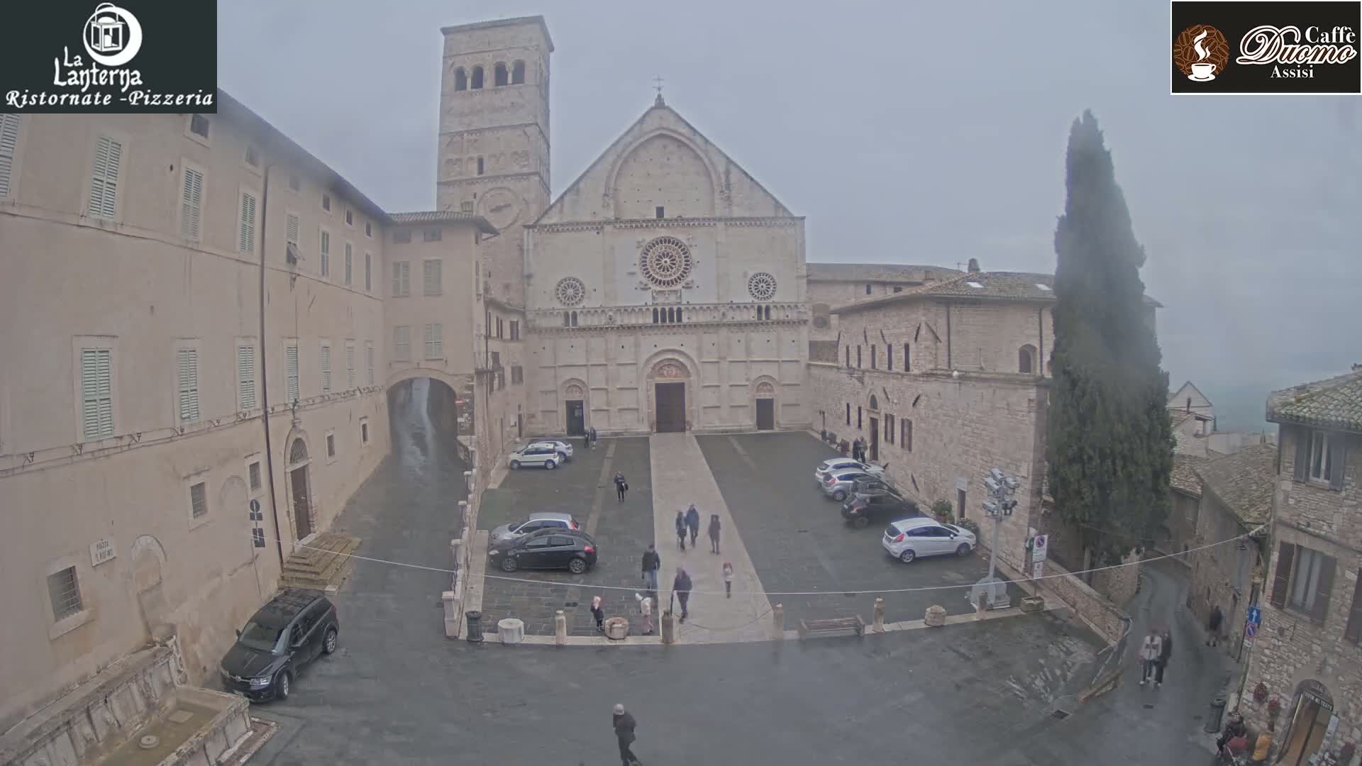 A large stone cathedral sits in a town square on an overcast day, with several cars parked and a few pedestrians walking around.
