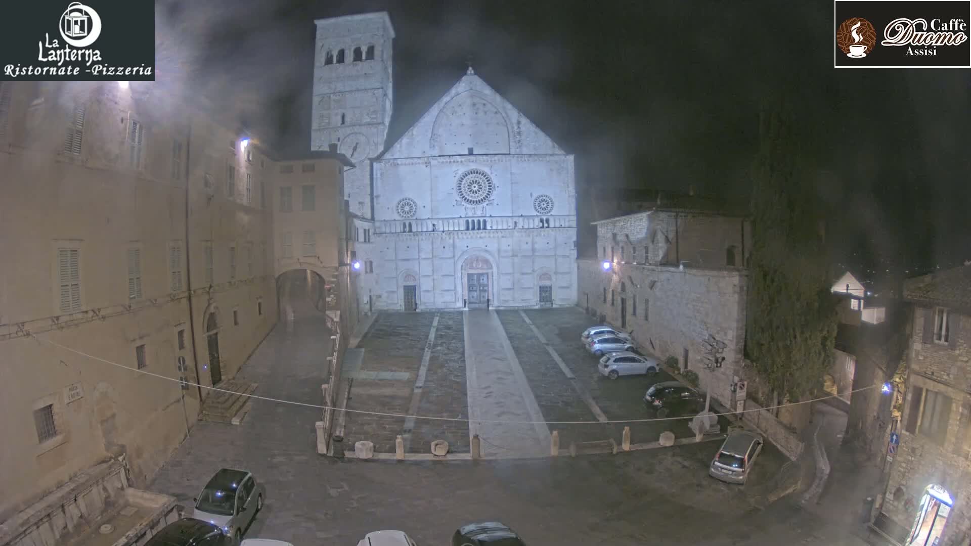 A large, light-colored cathedral stands in a town square at night, shrouded in a light fog, with several cars parked nearby.