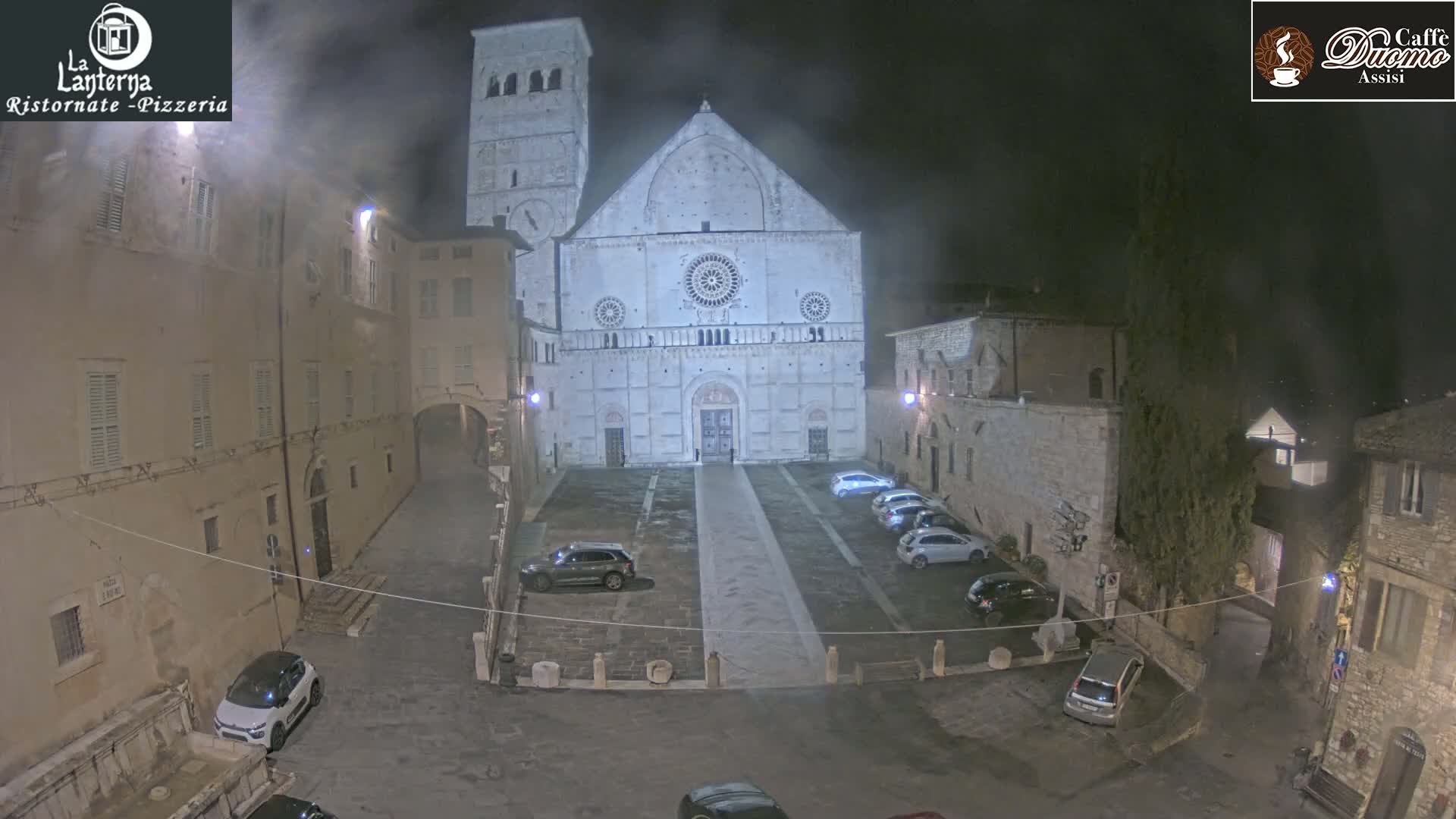 A nighttime view of a large, light-colored cathedral in a stone plaza with several parked cars, under hazy conditions.