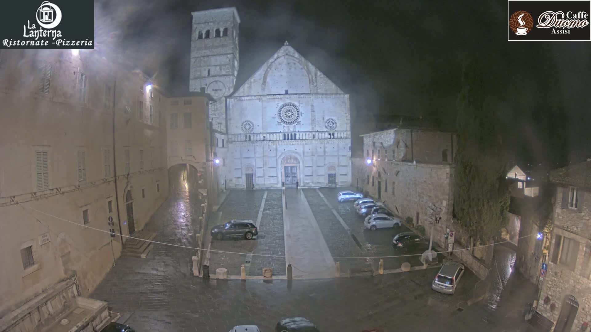 A large stone cathedral stands in a town square at night during light rain, with several cars parked around the square and buildings lining the sides.