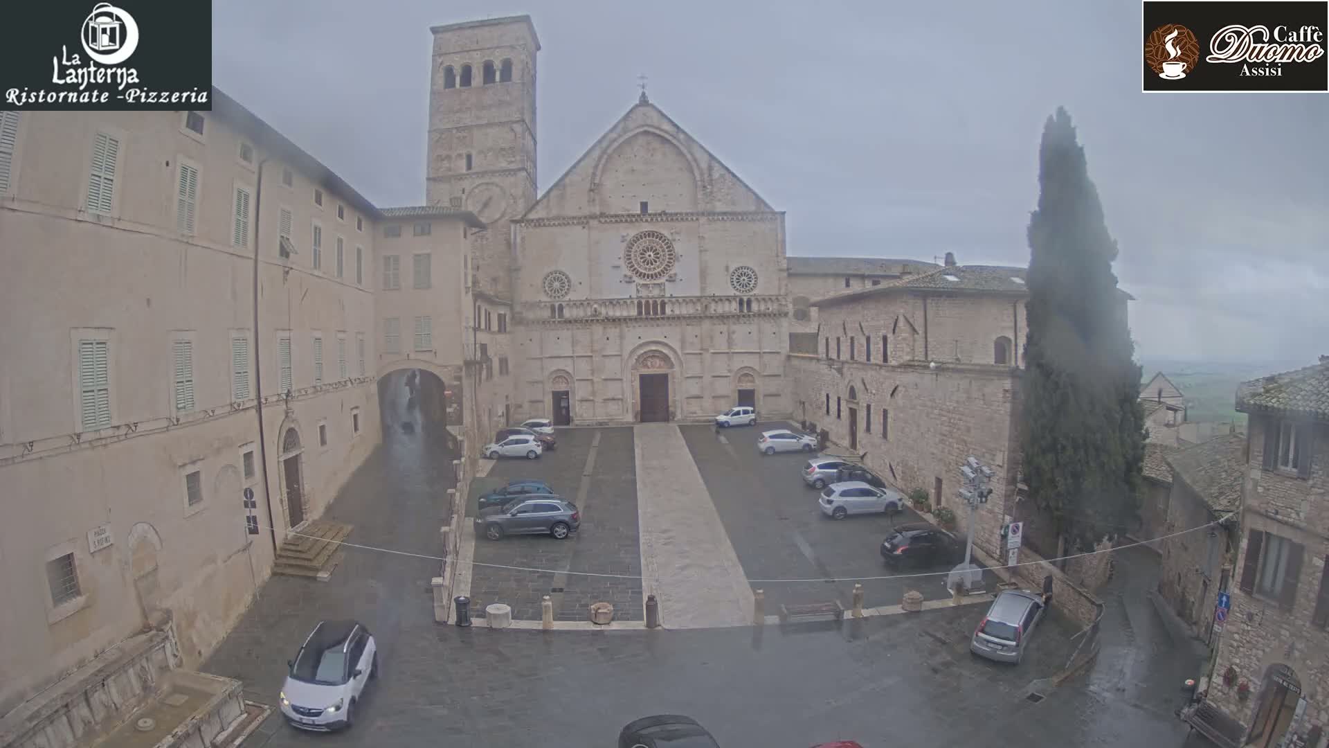 A large stone cathedral stands in a town square, surrounded by buildings, with cars parked and a light rain falling.