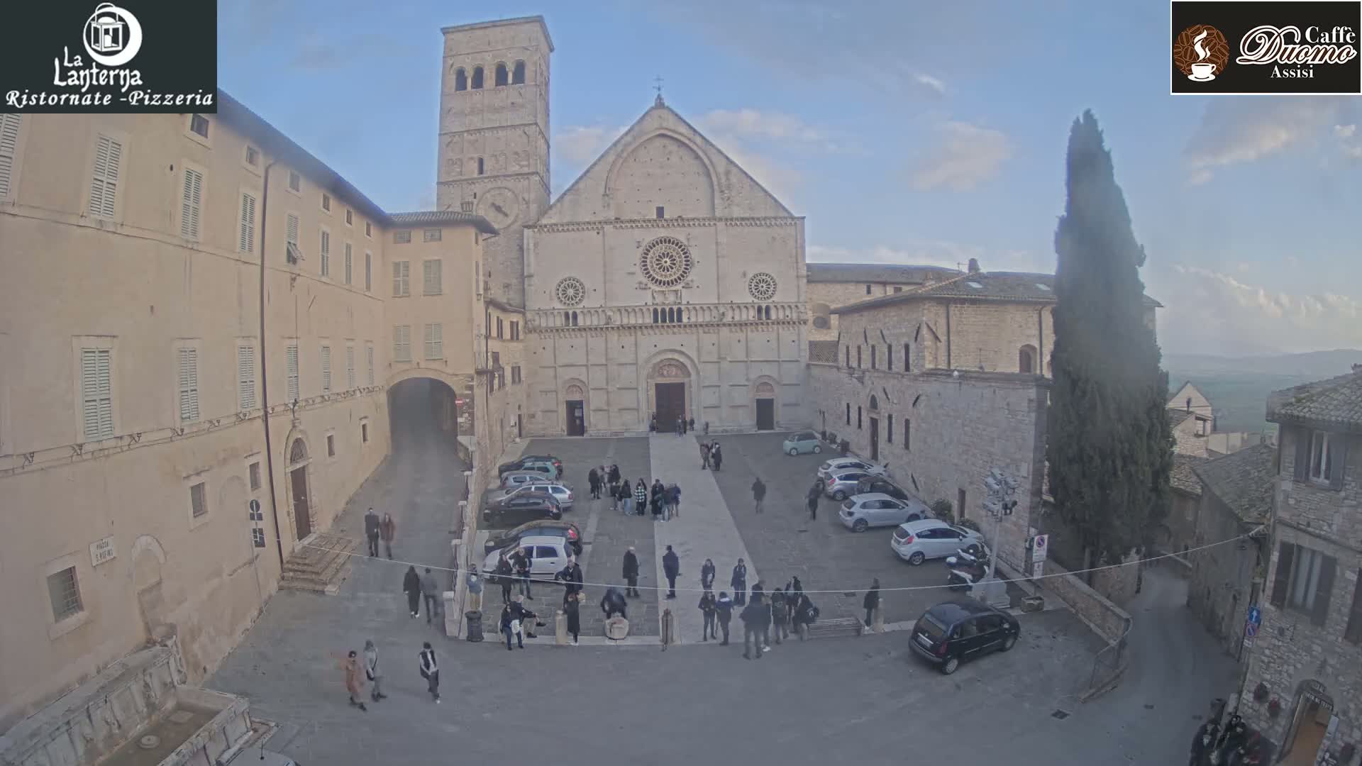 A large stone church stands in a town square, surrounded by buildings and a few parked cars, under a mostly clear, pale sky.