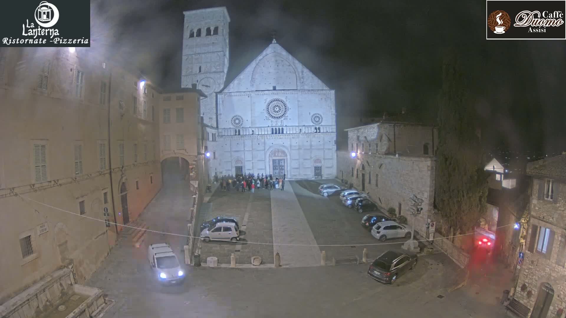 A nighttime view of a town square with a large cathedral, several cars parked, and a small group of people gathered in front of the cathedral, under a hazy, dark sky.