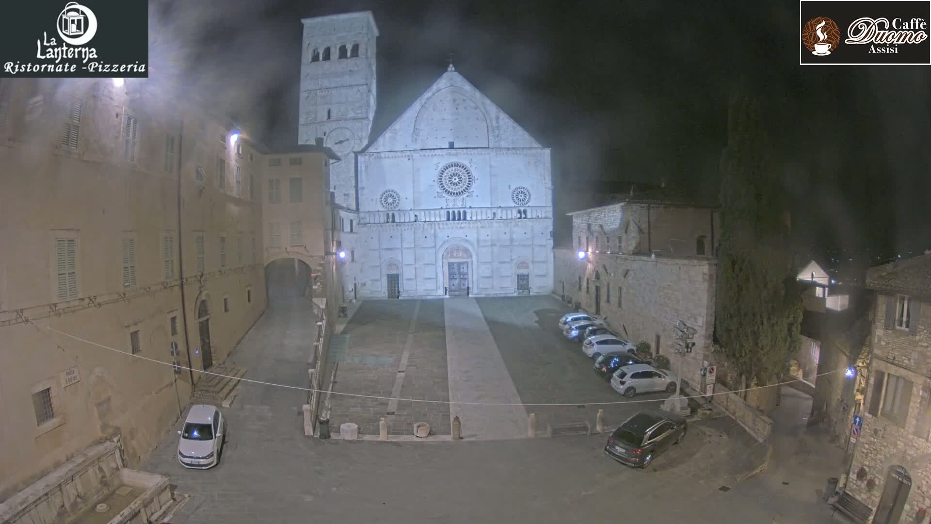 A large stone cathedral stands in a dimly lit, foggy town square at night, with several cars parked nearby.