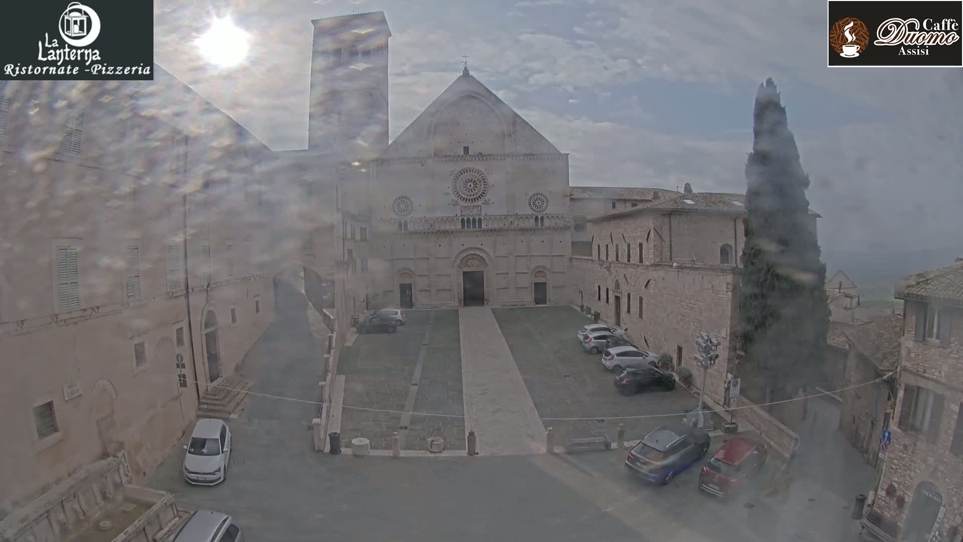 A hazy sun shines on a stone plaza with parked cars and buildings, one of which is a large, pale stone church.