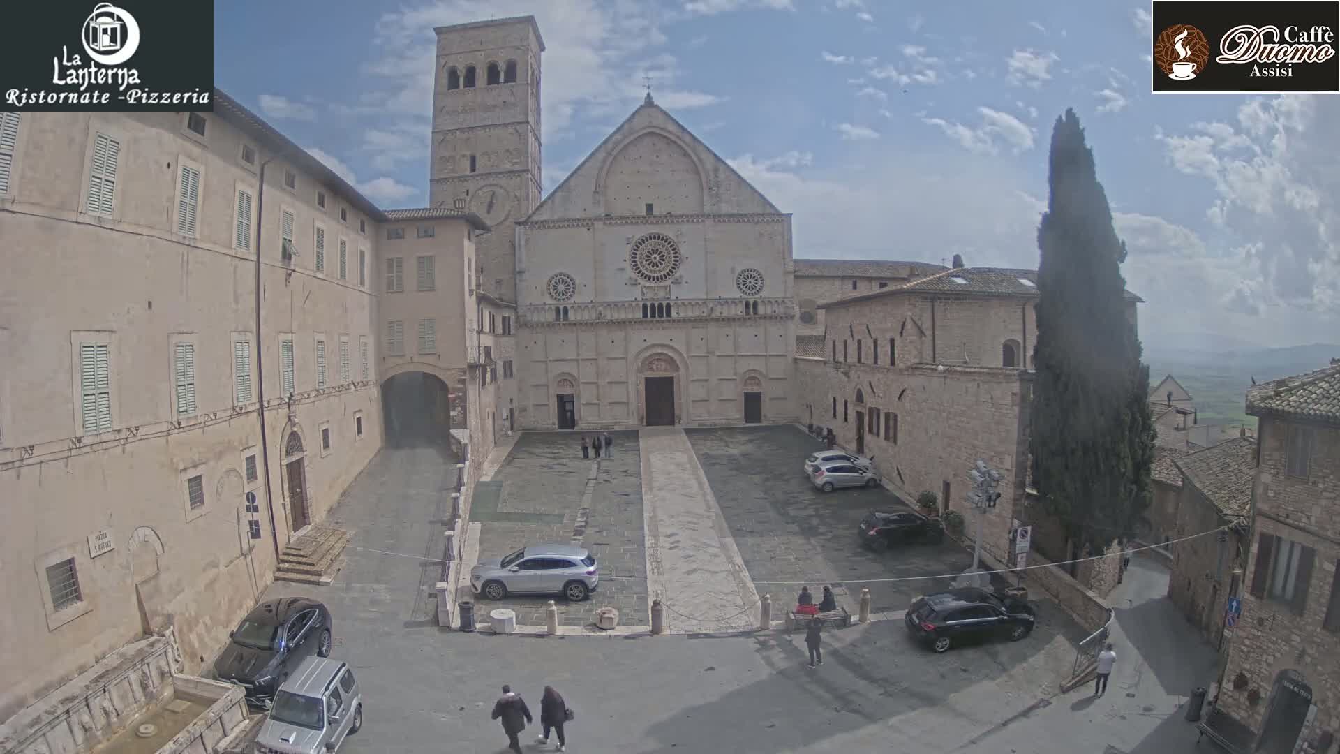 A large stone church is situated in a town square, surrounded by buildings, with a few cars parked and people walking, under a partly cloudy sky.