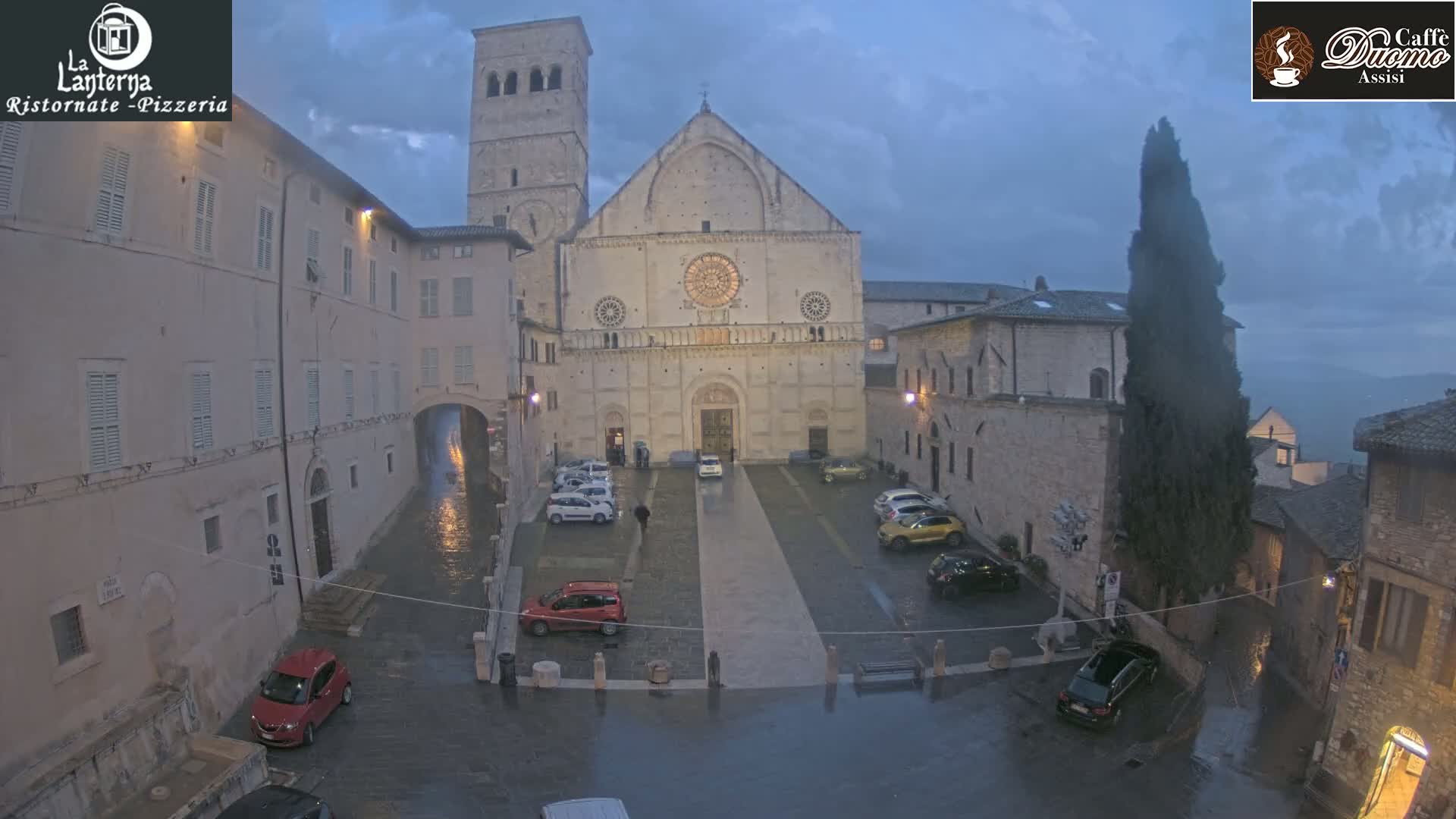 A large stone cathedral is situated in a town square, surrounded by buildings, with several cars parked, and a wet cobblestone surface indicating rain.
