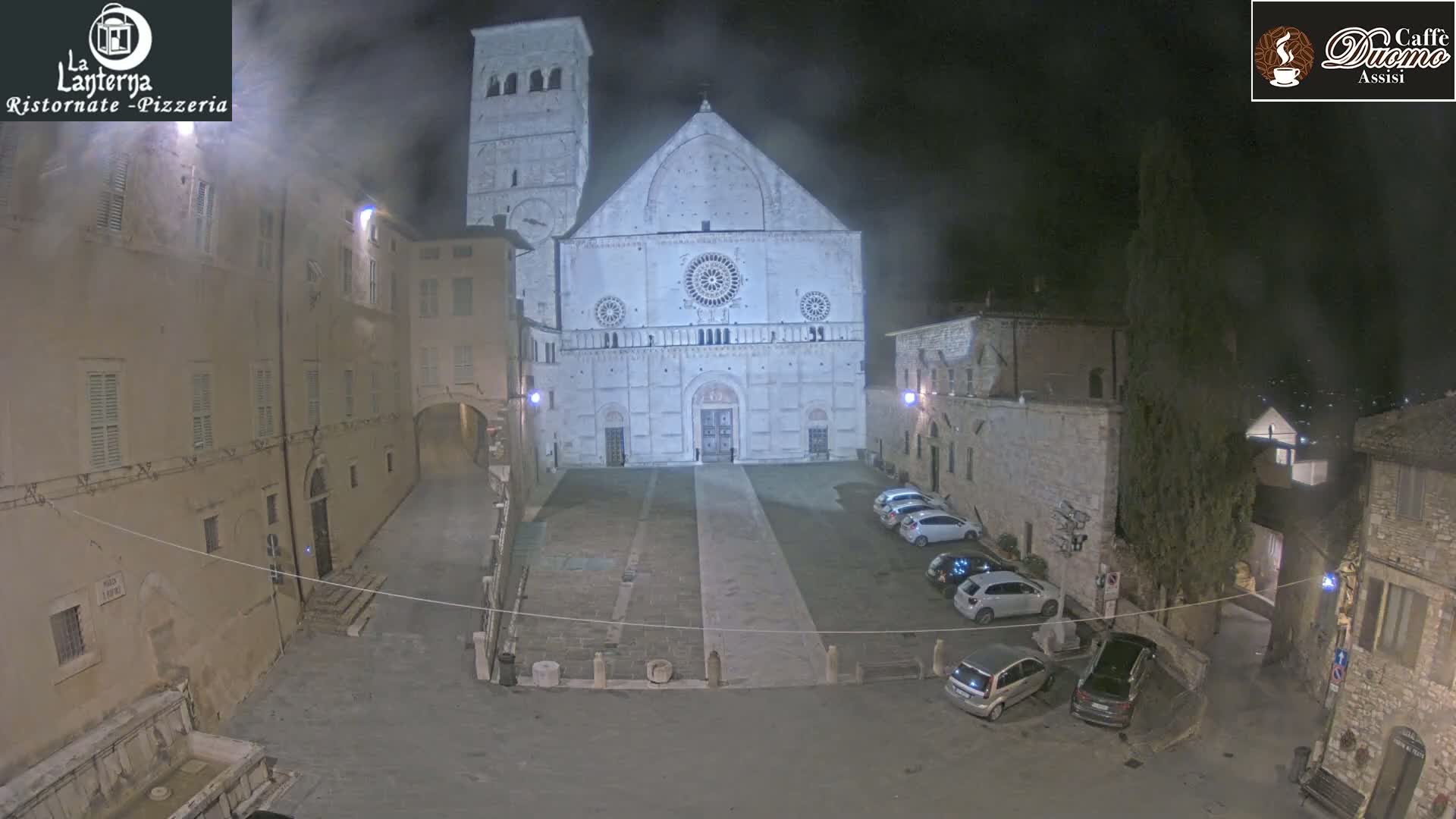 A nighttime view of a town square, featuring a large stone cathedral, several parked cars, and buildings surrounding a paved plaza under hazy or foggy conditions.