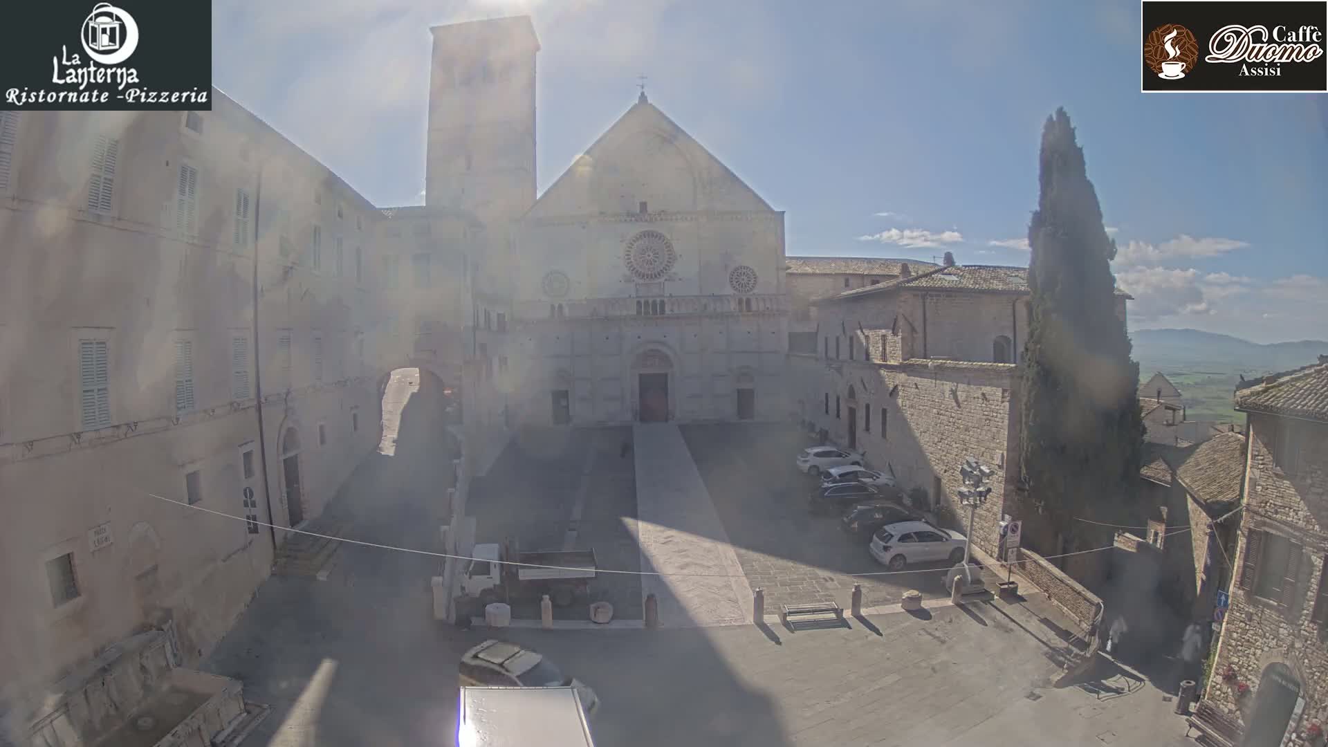 A sunny plaza with parked cars is situated in front of a large stone church, surrounded by other stone buildings.