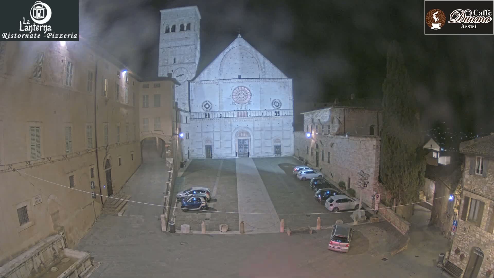 A nighttime, foggy view of a large, light-colored cathedral in a European-style town square with several cars parked and buildings surrounding the square.