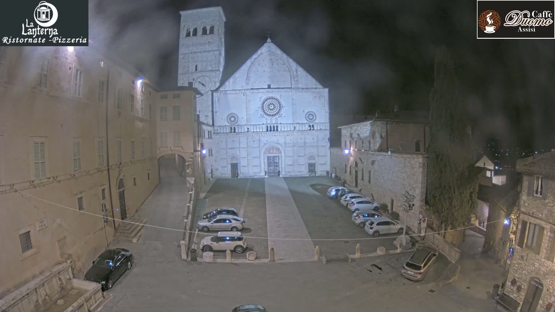 A nighttime view of a town square with several cars parked, centered on a large, light-colored cathedral under a hazy sky.