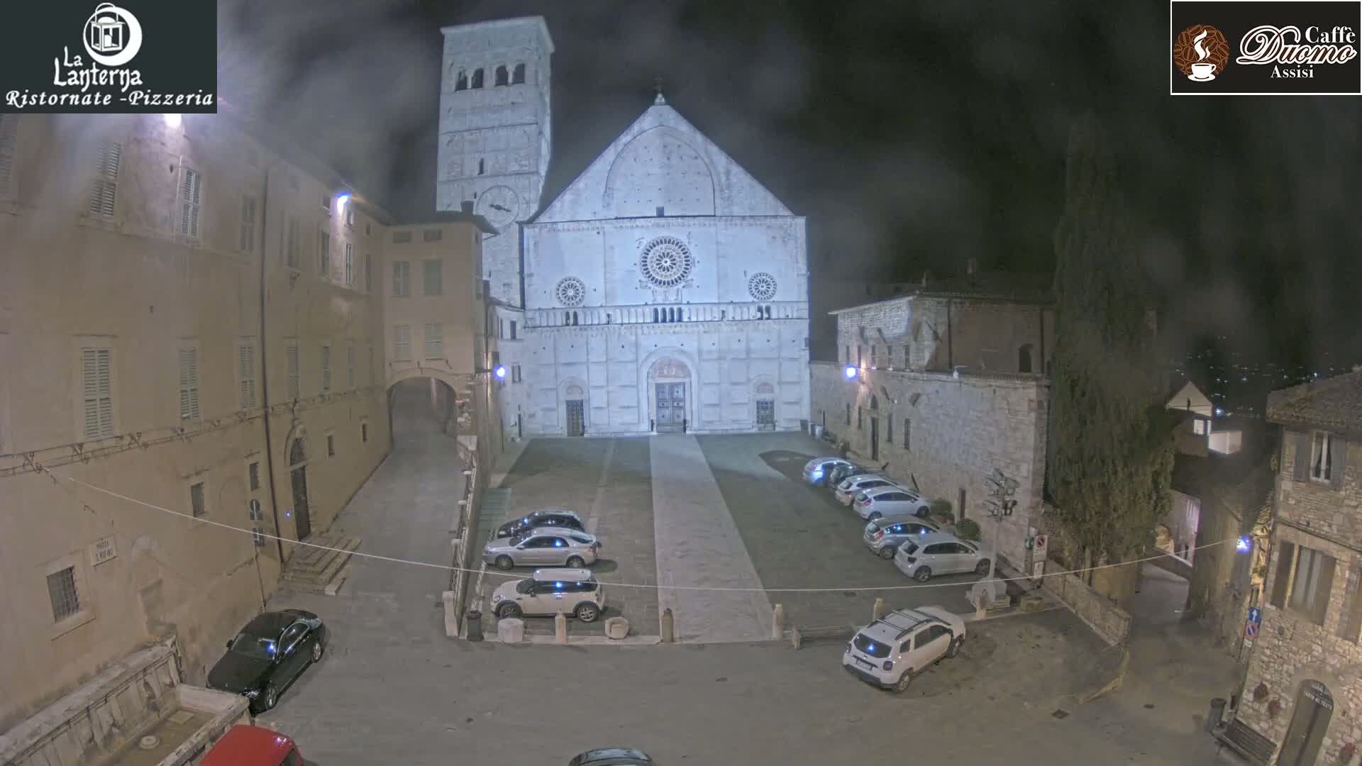 A nighttime, hazy view of a town square with several cars parked in front of a large, light-colored cathedral.