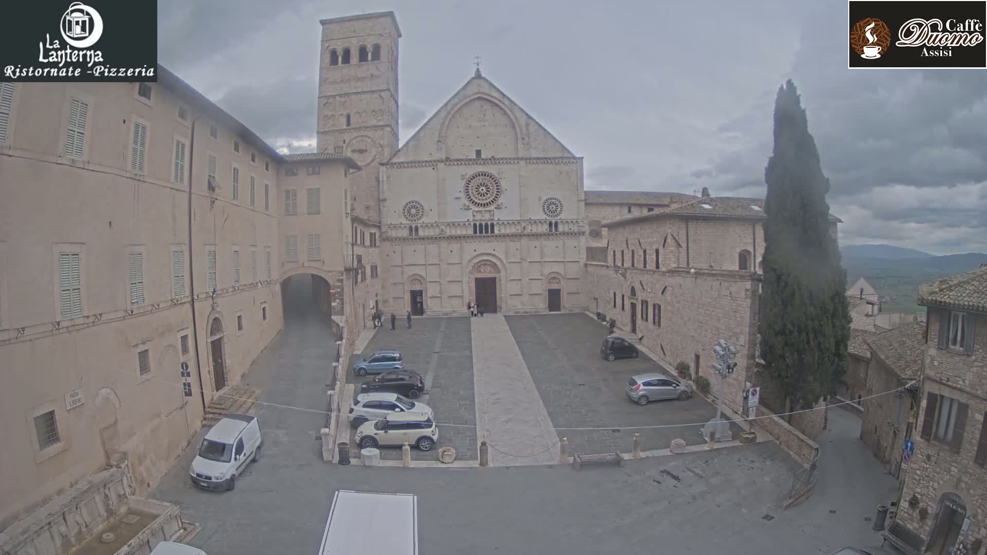 A stone cathedral stands in a town square on an overcast day, surrounded by several parked cars and older stone buildings.