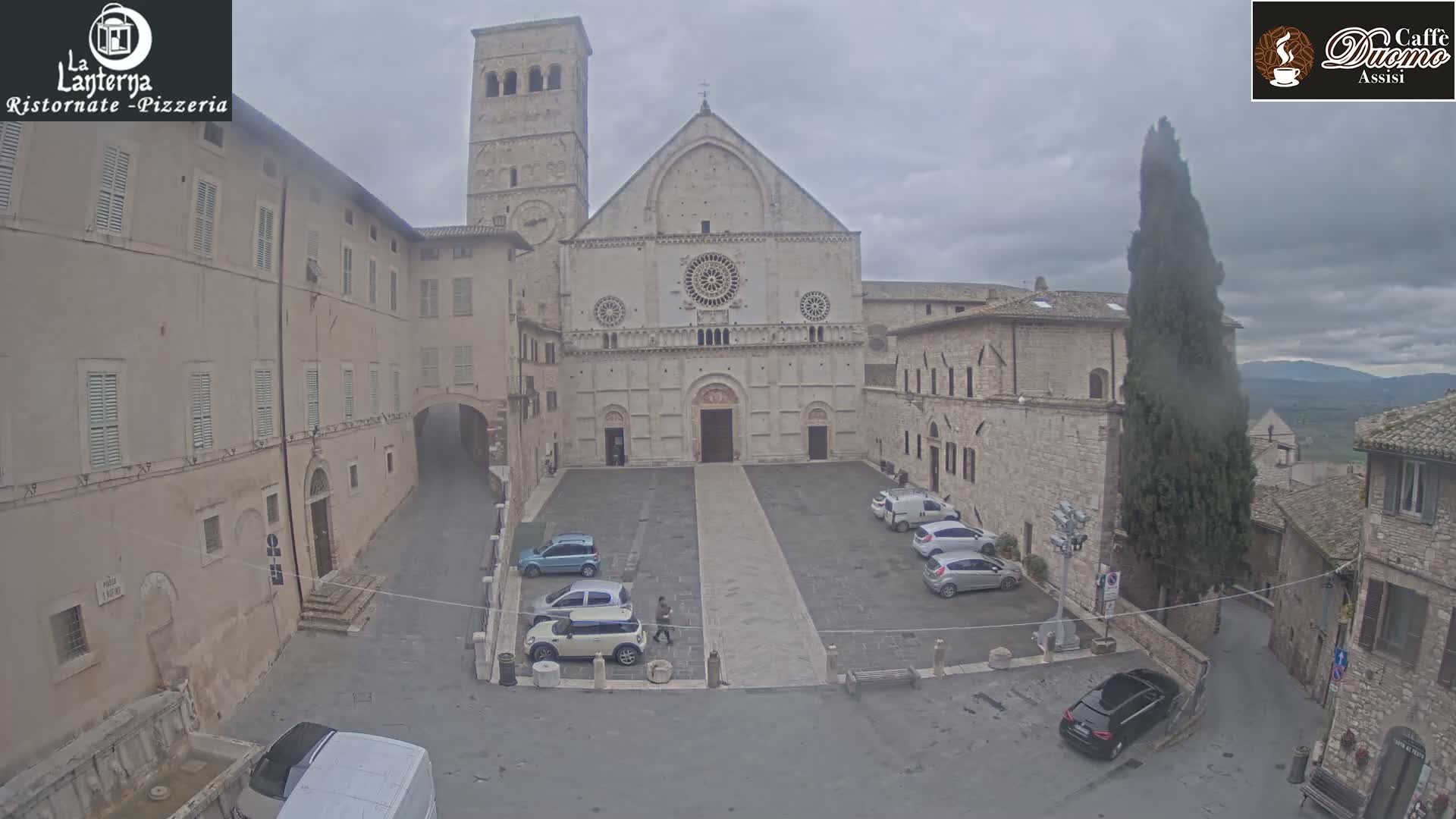 A large stone cathedral sits in a town square with several cars parked, all under an overcast sky.