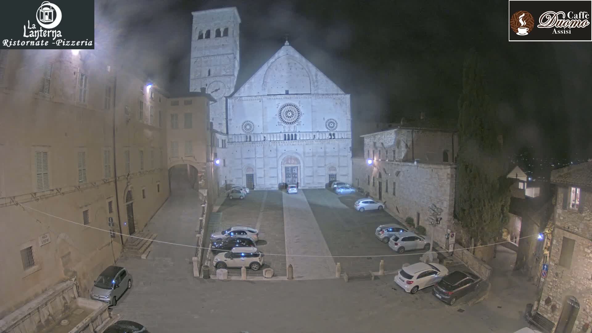 A nighttime view of a town square with parked cars in front of a large stone cathedral under a hazy sky.