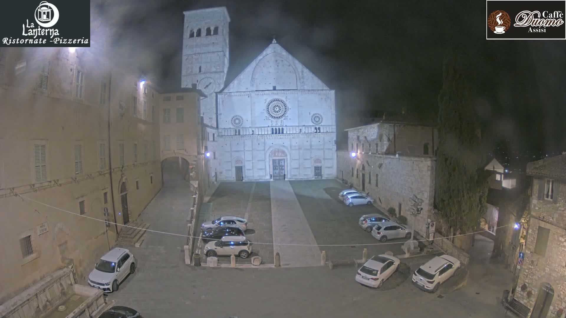 A nighttime view of a town square with parked cars in front of a large, light-colored cathedral under hazy conditions.
