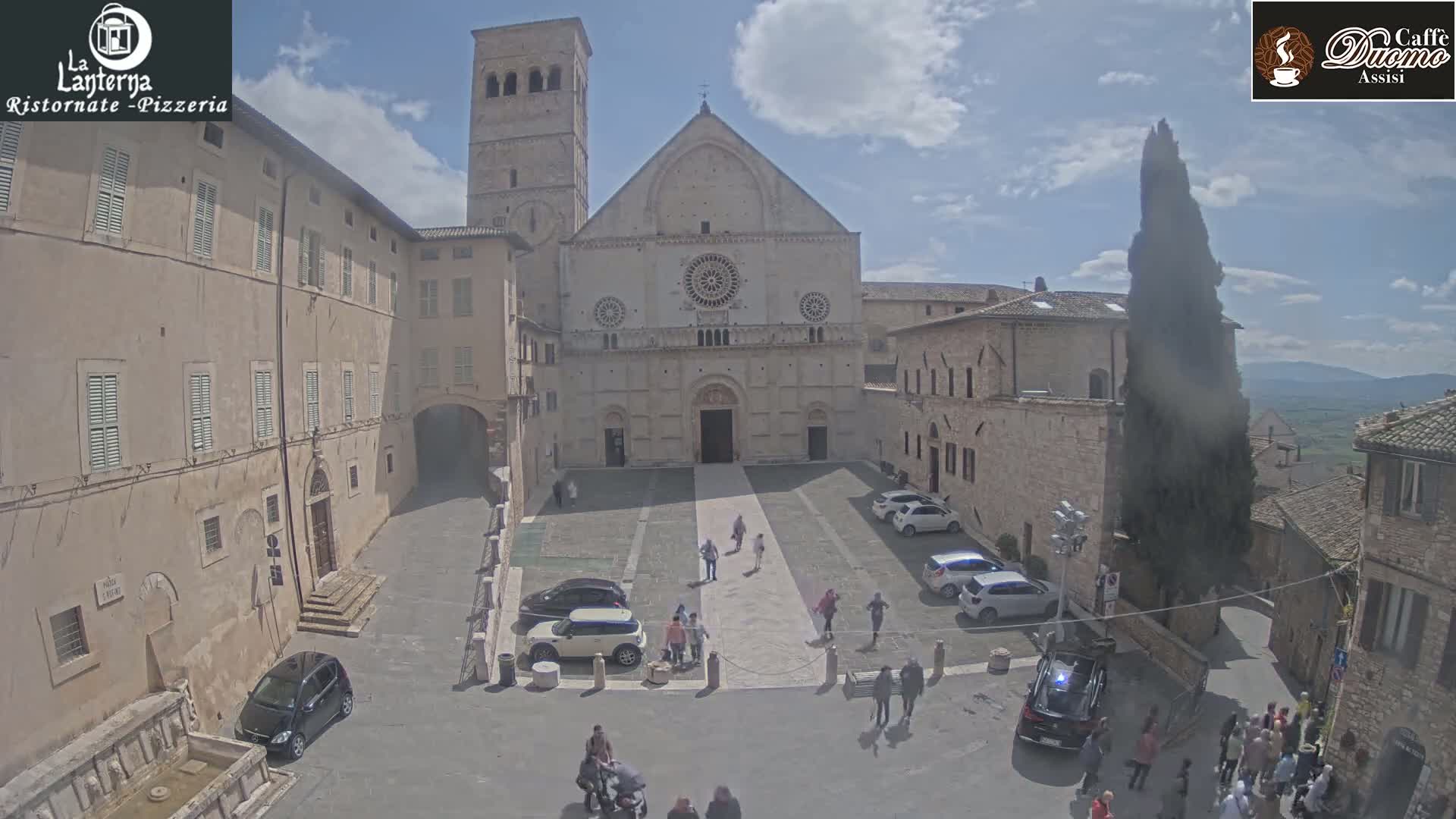 A large stone cathedral sits in a sunny town square, surrounded by other buildings and a few cars, with some people walking around.