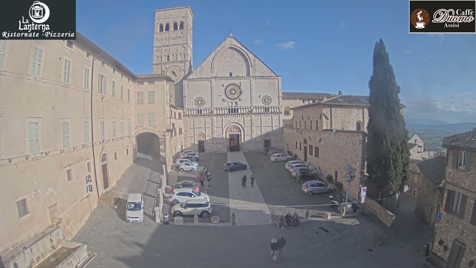 A large stone church sits in a sunny town square surrounded by buildings, cars, and a few pedestrians.