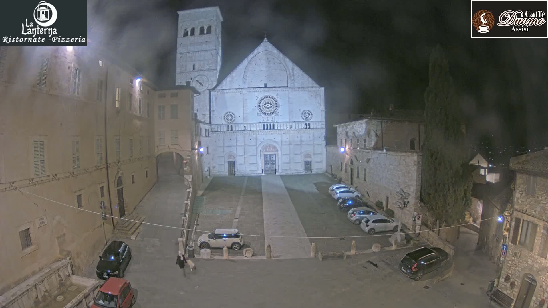 A nighttime, hazy view of a town square with a large cathedral in the background, several cars parked, and buildings lining the square.