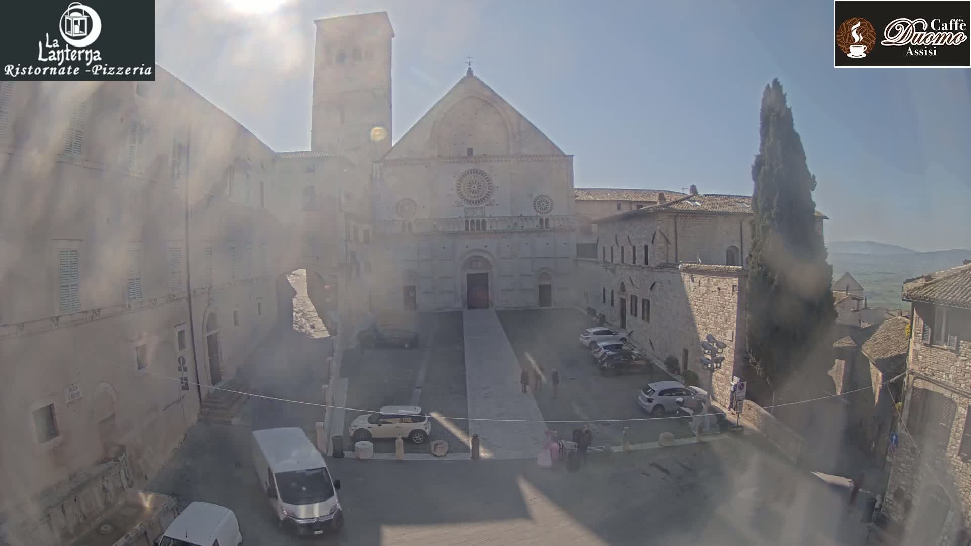 A sunny day reveals a large stone cathedral in a town square, surrounded by aged buildings and a few parked cars.