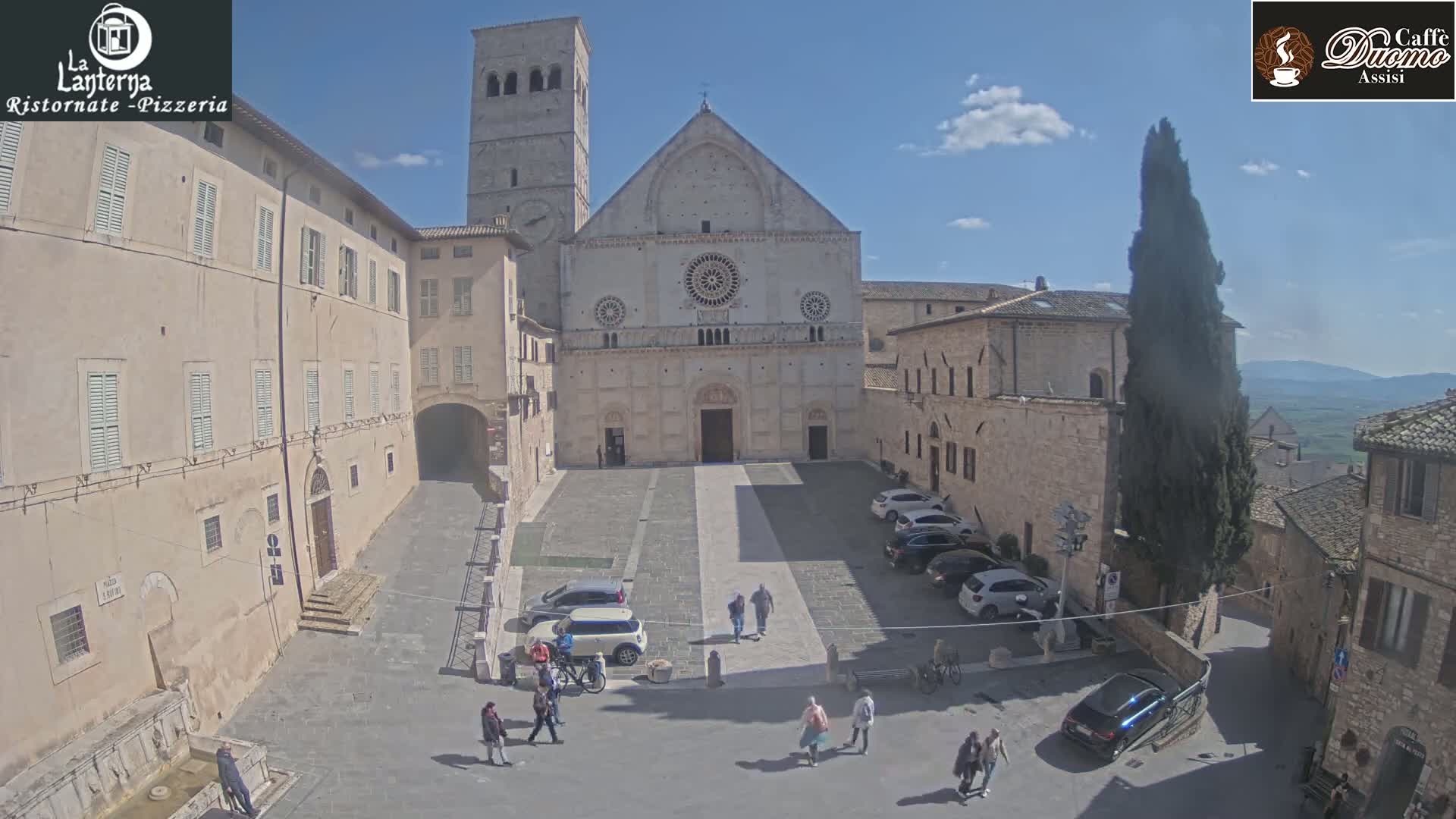 A large stone cathedral and several buildings surround a paved town square with a few parked cars and pedestrians on a sunny day.